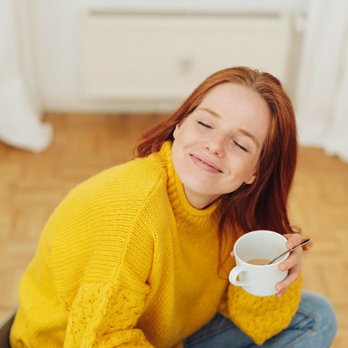 Young woman enjoying a cup of tea seated cross-legged on the floor looking up at the camera with closed eyes and a smile